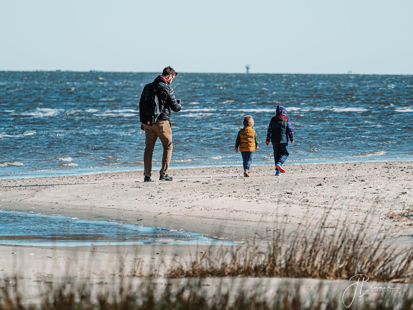 A man and two young children bundled in jackets walk along the shoreline of Tybee Island on a clear winter day.