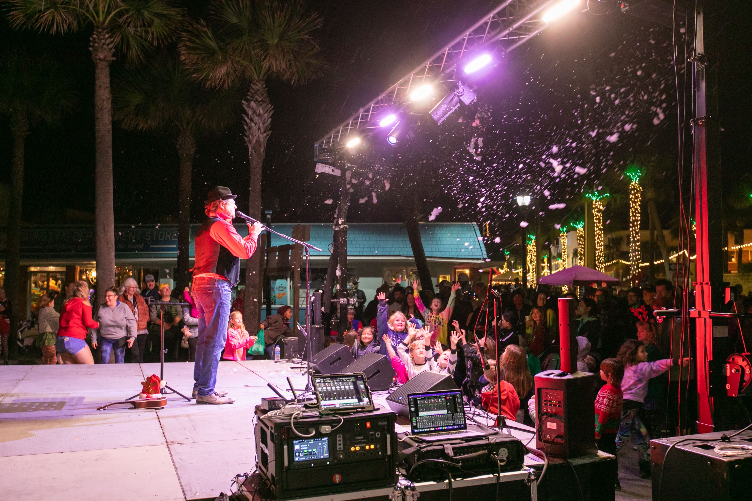 A performer entertains families gathered around an outdoor stage during Tybee Holly Days, as artificial snow falls and palm trees glow with holiday lights.
