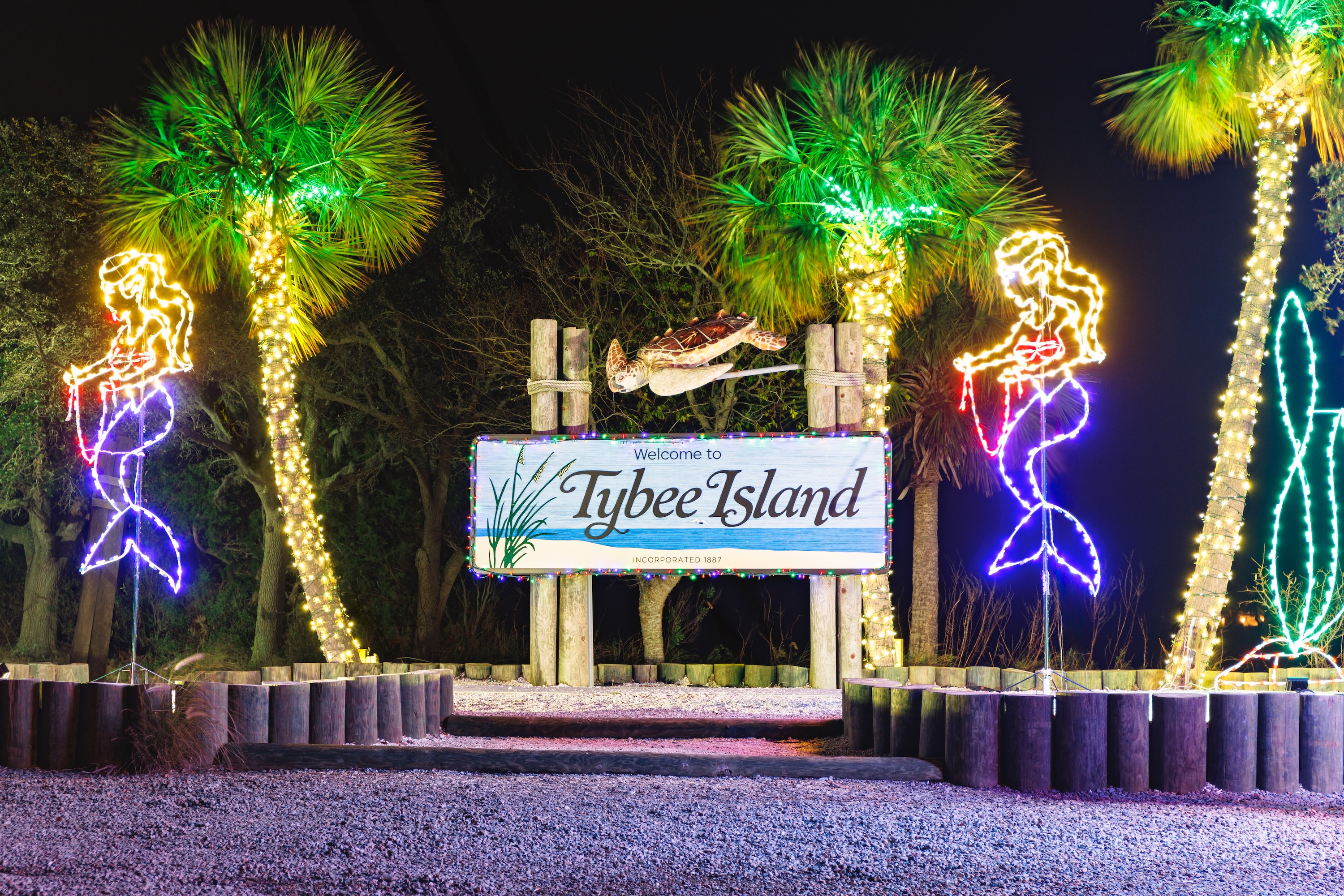 A Welcome to Tybee Island sign surrounded by illuminated palm trees, colorful holiday lights and glowing mermaid decorations at night.