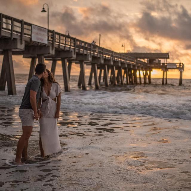 Couple kissing on the beach at sunset with a pier in the background