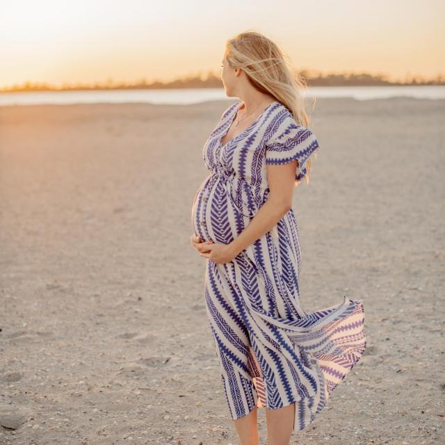 A woman standing on the beach in a dress holding her pregnant belly.