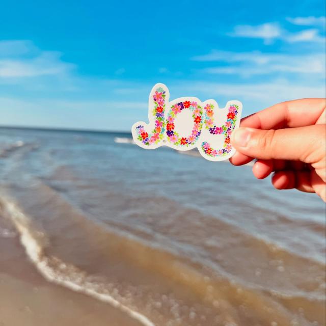 A person’s hand holding a sticker on the beach that reads: “Joy.”