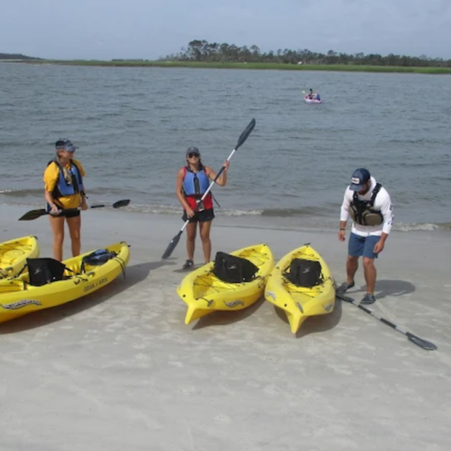 Three people stand on a sandy shoreline beside bright yellow kayaks, preparing for a guided kayaking excursion on calm coastal waters.