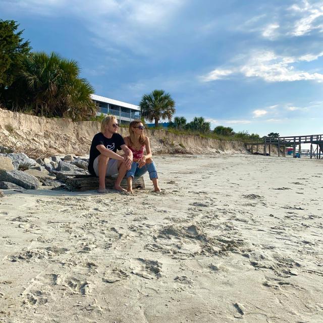 Two people sitting barefoot on the sand along Tybee Island’s shoreline, with dunes, palm trees and a pier in the distance under a blue sky.