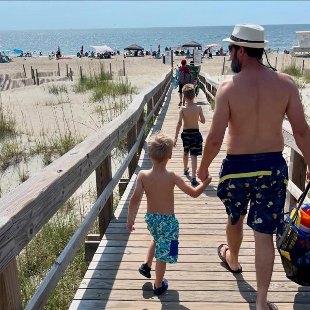 A father holds his son’s hand while walking towards the beach.