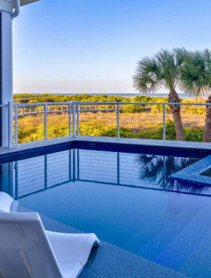 Private pool with in-water lounge chairs overlooking coastal dunes and palm trees on Tybee Island, with a screened porch and elevated deck seating.