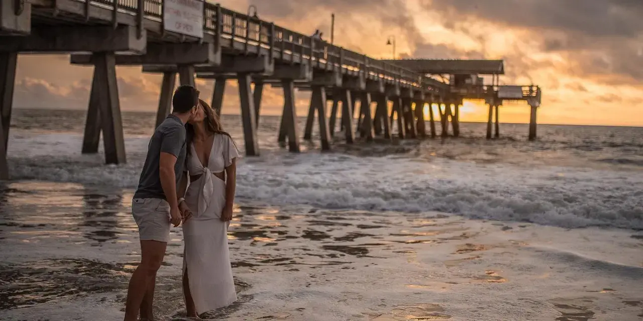 Couple kissing on the beach at sunset with a pier in the background