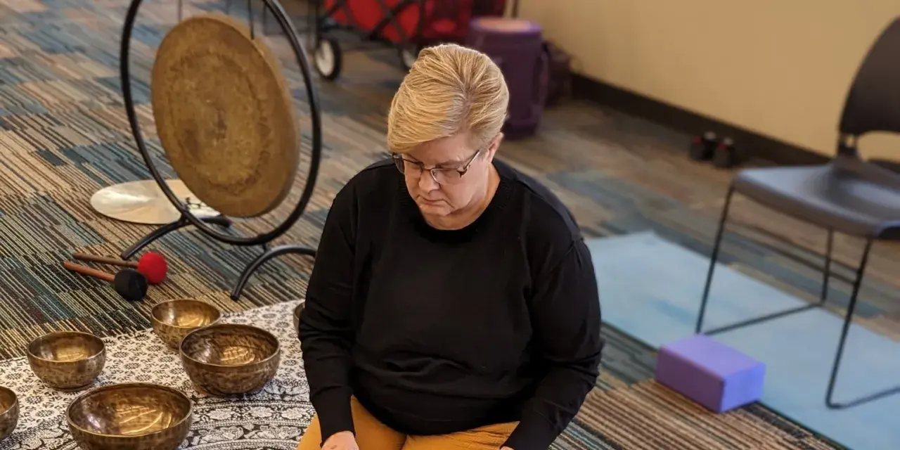 Woman seated on a patterned mat playing a handpan during a sound healing session, surrounded by singing bowls, a gong and percussion instruments.