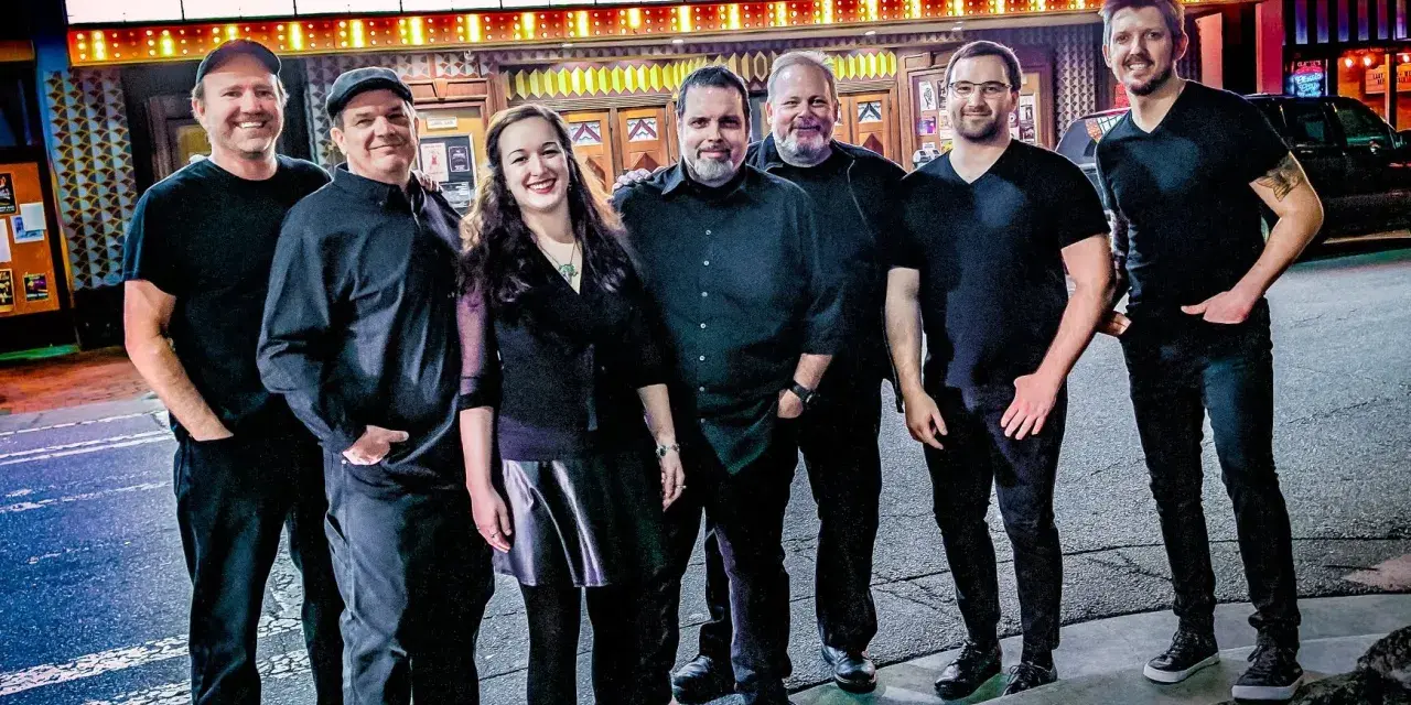 Seven members of Interstellar Echoes: A Tribute to Pink Floyd stand together and smile in front of a theater’s illuminated marquee.