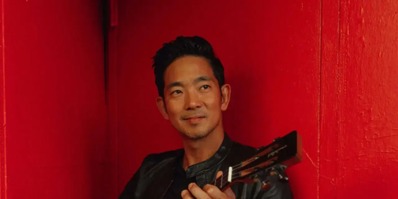 A ukulele artist is posing in front of a red wall.