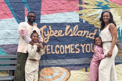 A family of four posing together in front of a wall mural sign that reads: Welcome to Tybee Island.