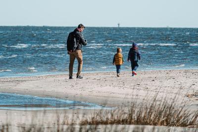 A man and two young children bundled in jackets walk along the shoreline of Tybee Island on a clear winter day.
