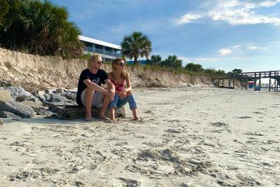 Two people sitting barefoot on the sand along Tybee Island’s shoreline, with dunes, palm trees and a pier in the distance under a blue sky.