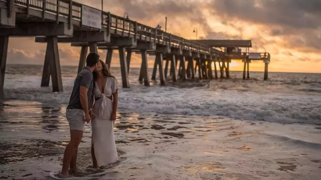 Couple kissing on the beach at sunset with a pier in the background