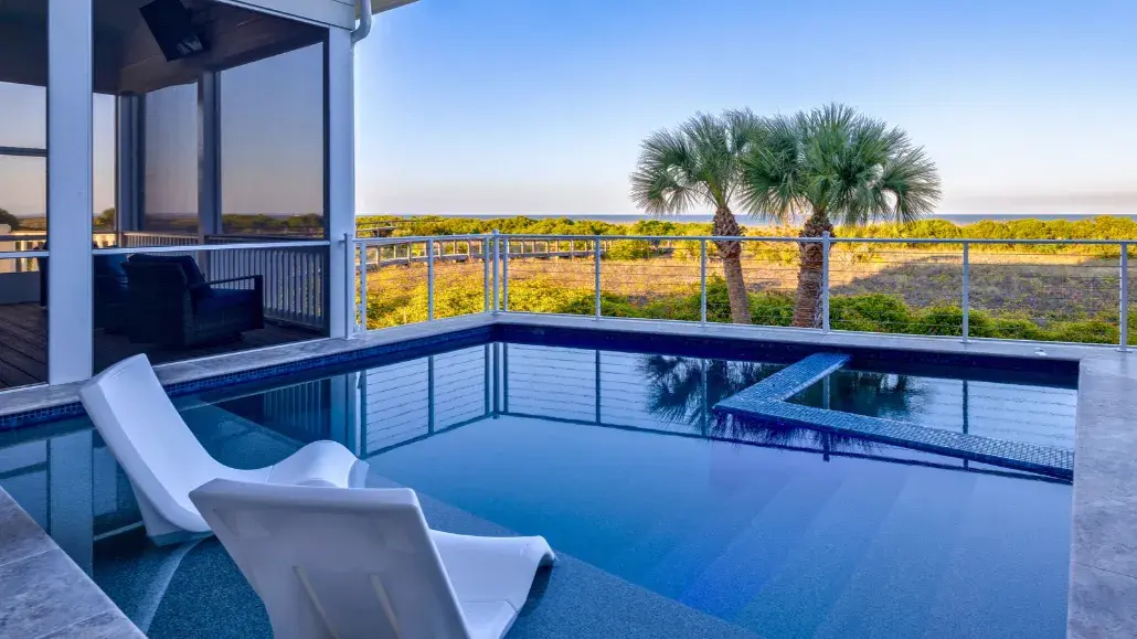 Private pool with in-water lounge chairs overlooking coastal dunes and palm trees on Tybee Island, with a screened porch and elevated deck seating.