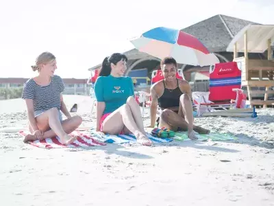 A girl’s group of three sitting and laughing on the beach on a sunny day.
