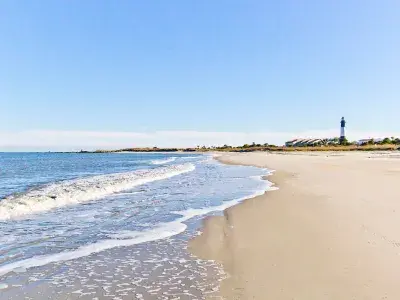 Wide view of a sandy beach with gentle waves rolling in and the Tybee Island Lighthouse visible in the distance.