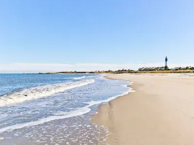 Wide view of a sandy beach with gentle waves rolling in and the Tybee Island Lighthouse visible in the distance.