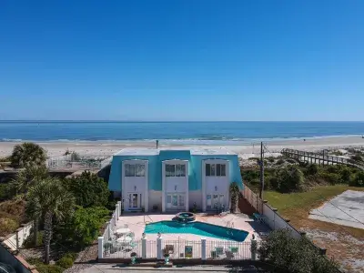 Aerial backyard view of a blue and white beach home’s pool with the beach in the foreground.