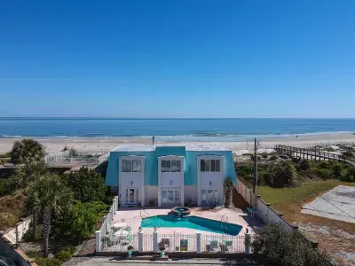 Aerial backyard view of a blue and white beach home’s pool with the beach in the foreground.