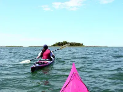 View from a red kayak of another kayaker paddling across open water toward a small island.