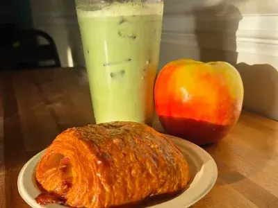 A croissant on a plate along with a iced mocha and apple.