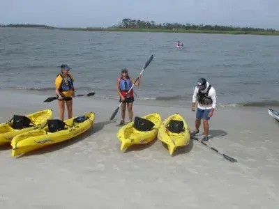 Three people stand on a sandy shoreline beside bright yellow kayaks, preparing for a guided kayaking excursion on calm coastal waters.