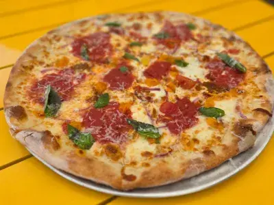 Wood-fired pizza topped with melted mozzarella, pepperoni, fresh basil and chili oil, served on a metal tray atop a bright yellow table.