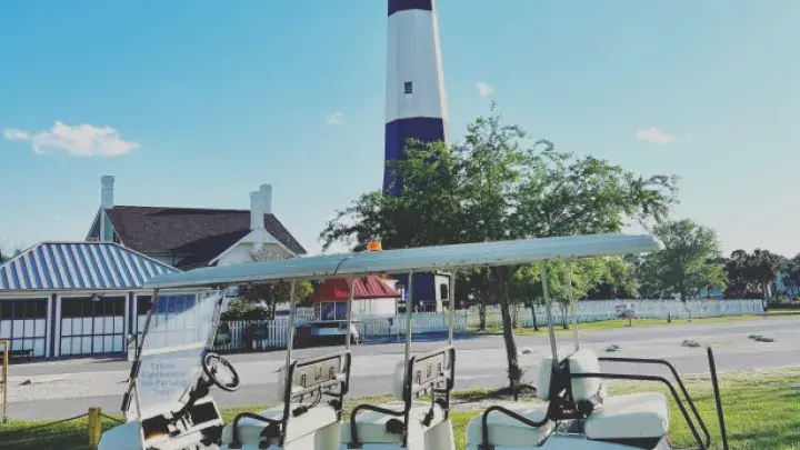 A multi-seat white golf cart parked on gravel with a lighthouse in the background.