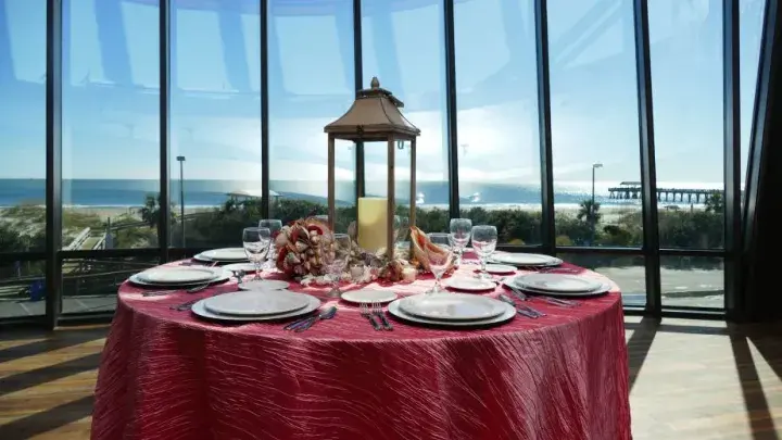 A table set with a red tablecloth and white dishes overlooking floor to ceiling windows on the beach.