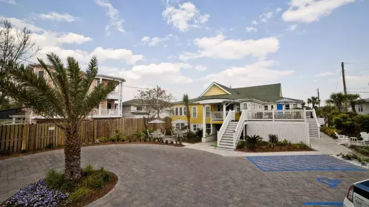 A charming yellow beach house with white stairs and a spacious deck, surrounded by tropical landscaping and a paved parking area.