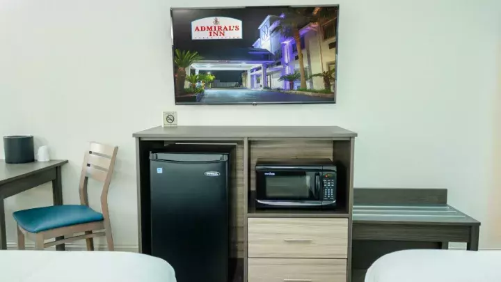 Hotel room with a mini-fridge, microwave, and a wall-mounted TV displaying the Admiral’s Inn logo and exterior.