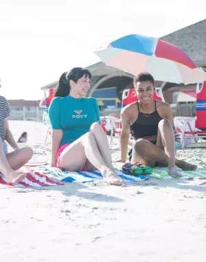 A girl’s group of three sitting and laughing on the beach on a sunny day.